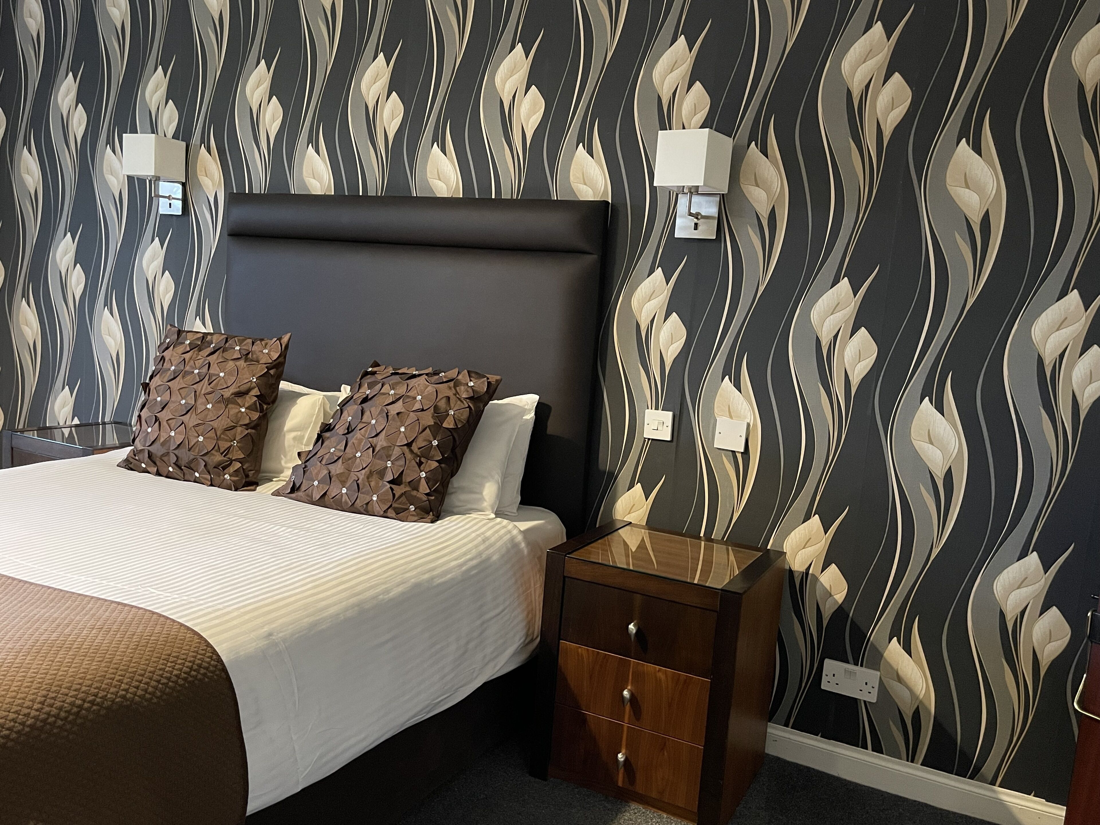 Un dormitorio con cama, dos almohadas, una mesita de noche con cajones y una pared con diseño de hojas.