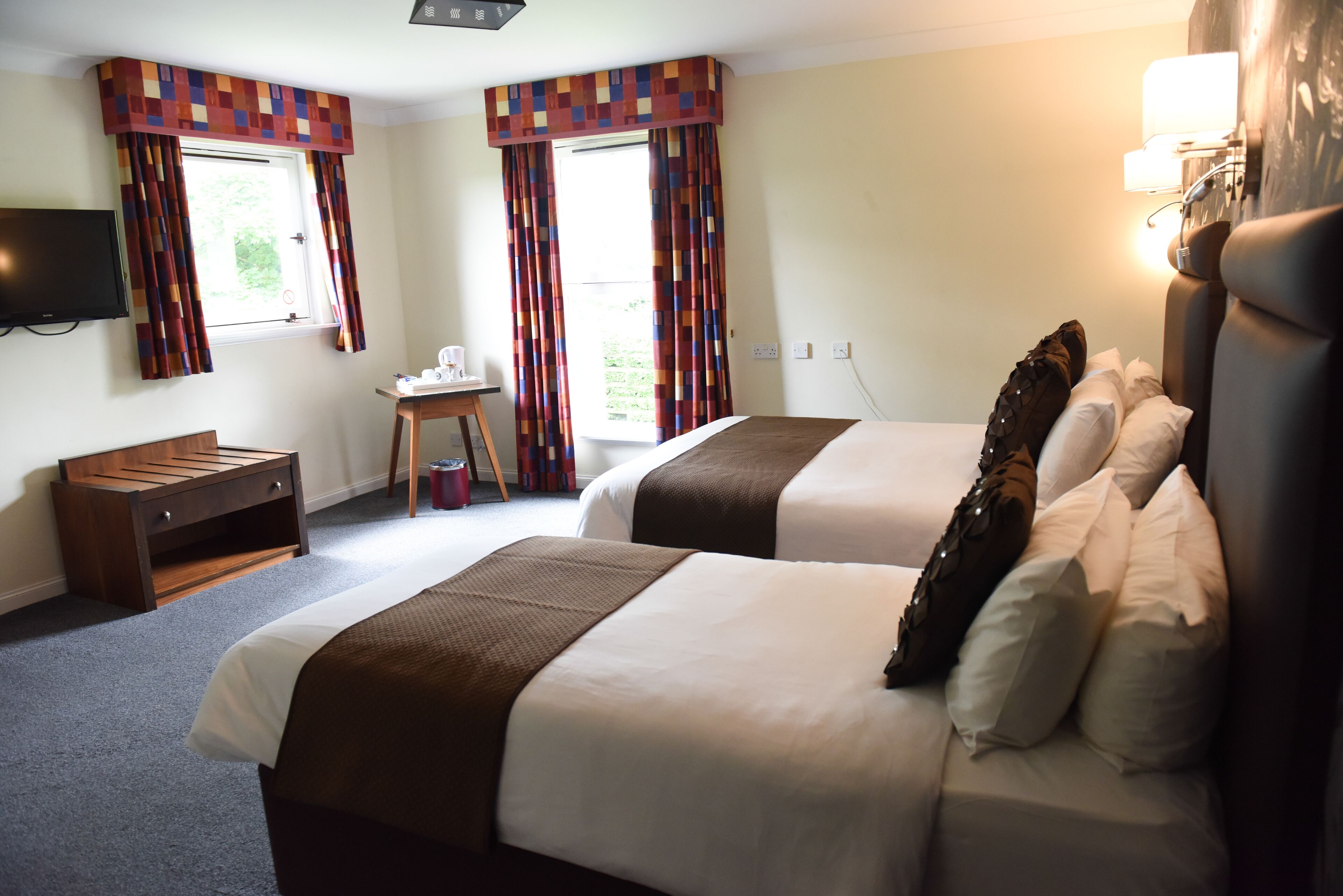 A hotel room with two beds, a TV, a small table, and a window with curtains.