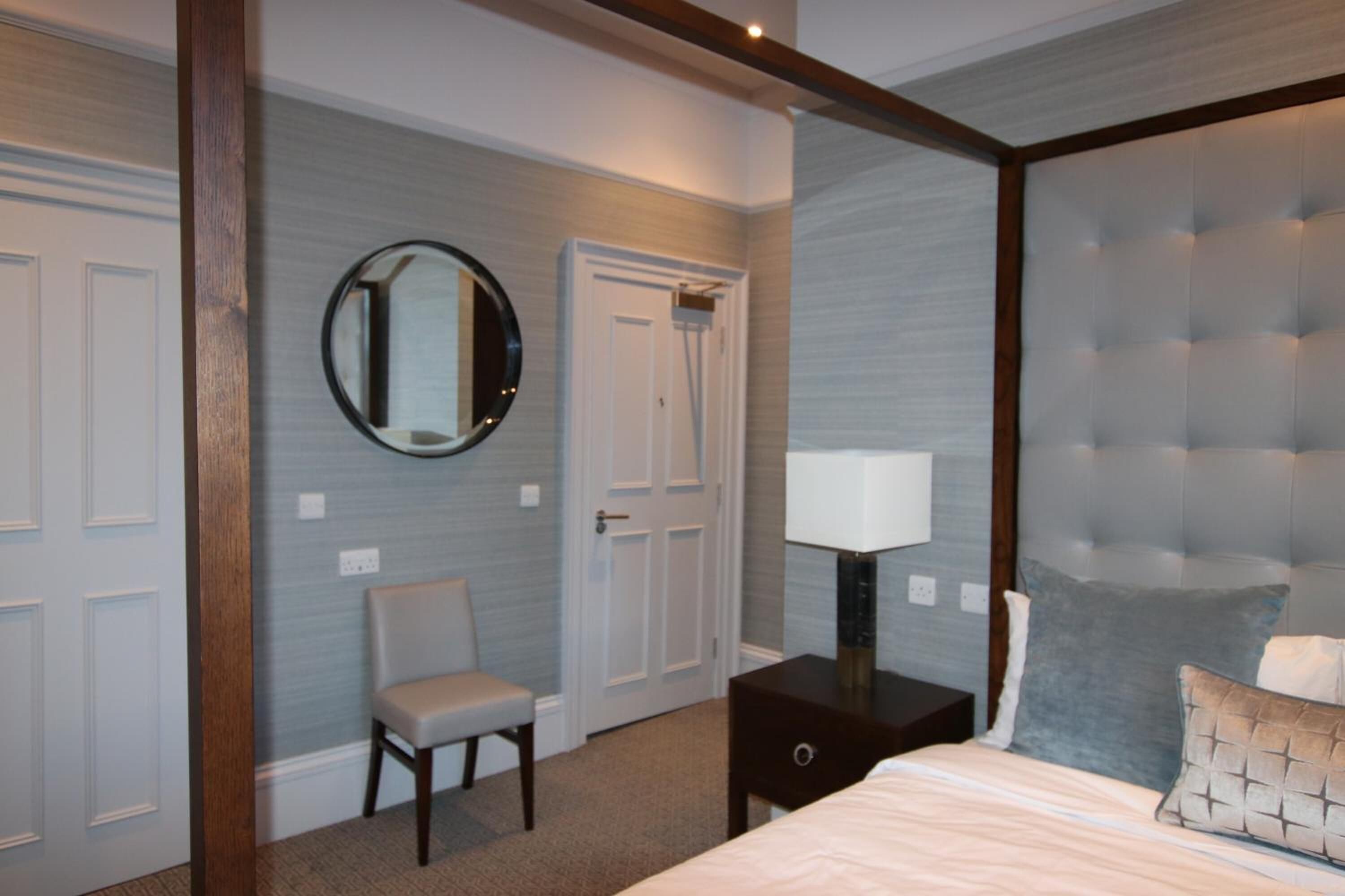 Un dormitorio con cama, una silla, una mesita de noche, una lámpara, un espejo y una puerta.