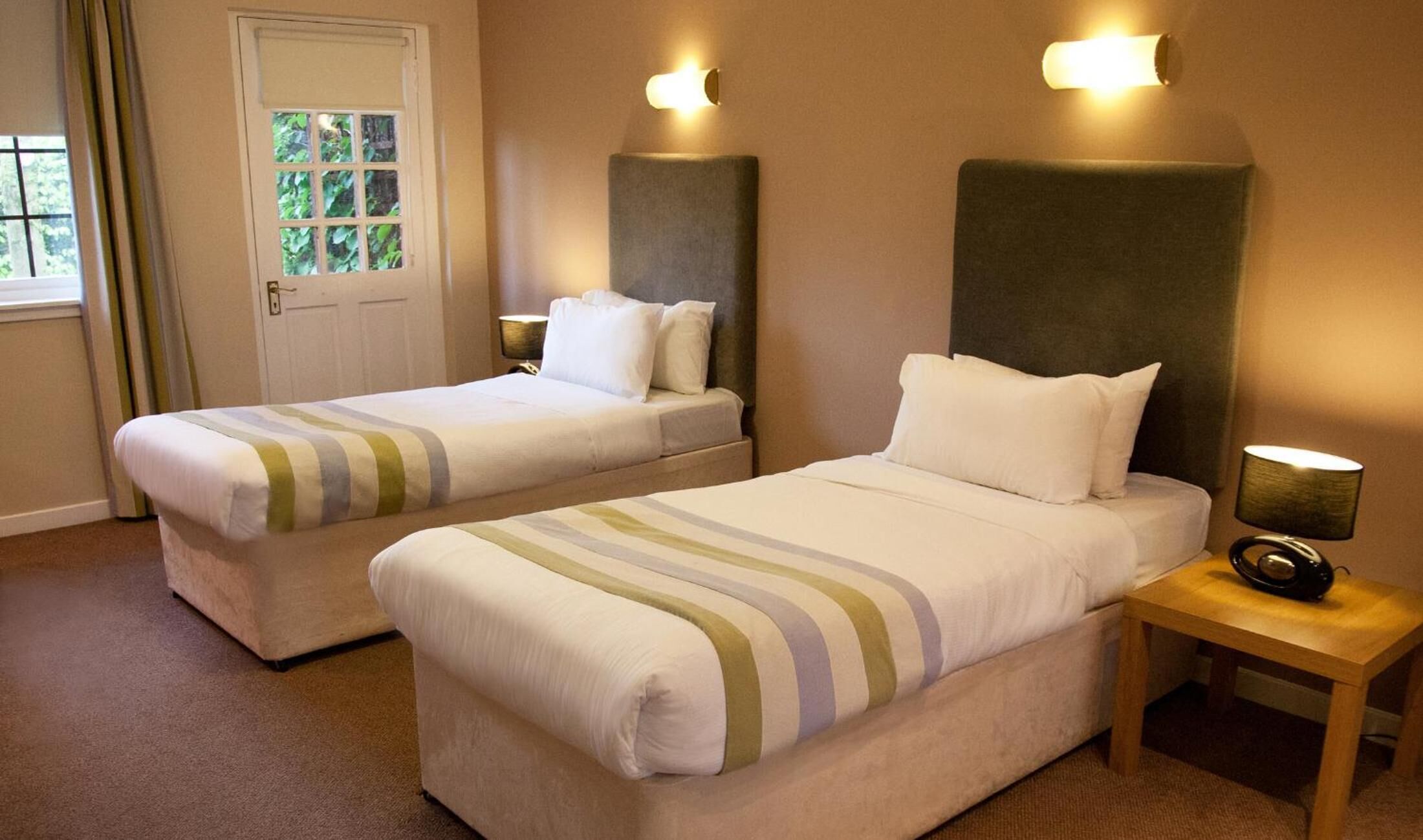 A hotel room with two single beds, a wooden nightstand, a lamp, and a door with a view of greenery outside.