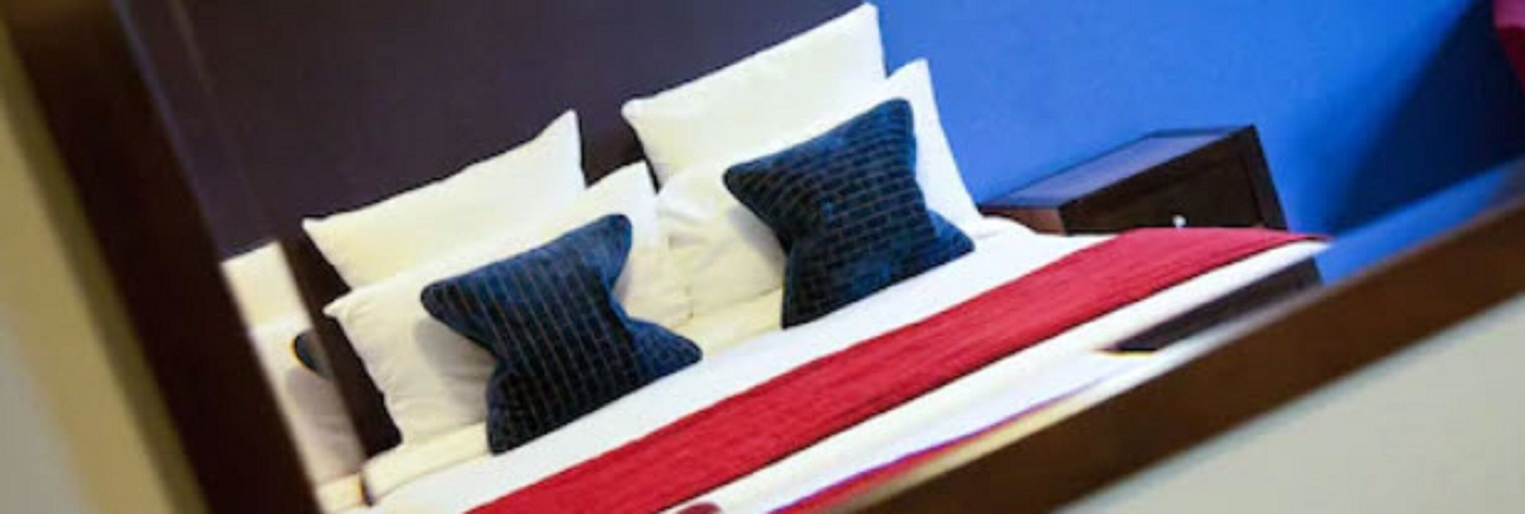 A bed with white and dark blue pillows, a red blanket, and a wooden nightstand with a lamp.