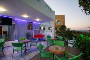 A rooftop terrace with green wicker furniture, a red cushioned chair, and a flat-screen TV. - Romantica Hotel Apartments (Hersonissos)