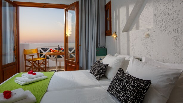 Une chambre d’hôtel avec un lit, une serviette verte, une chaise en bois, une table avec des fruits et une vue sur le coucher de soleil.