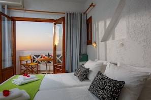 Ein Hotelzimmer mit Bett, einem grünen Handtuch, einem Holzsessel, einem Tisch mit Früchten und Blick auf den Sonnenuntergang.