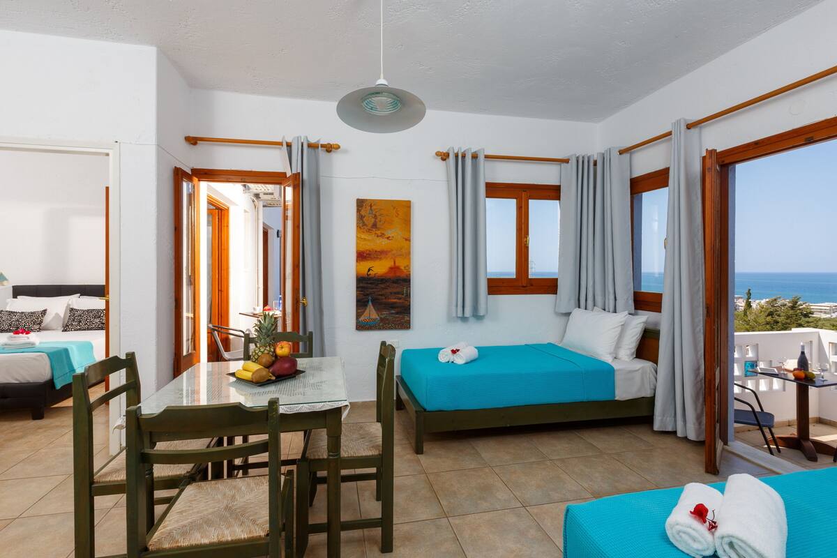 Een kamer met een bed, een eettafel met stoelen, een bijzettafeltje met fruit, en uitzicht op zee.