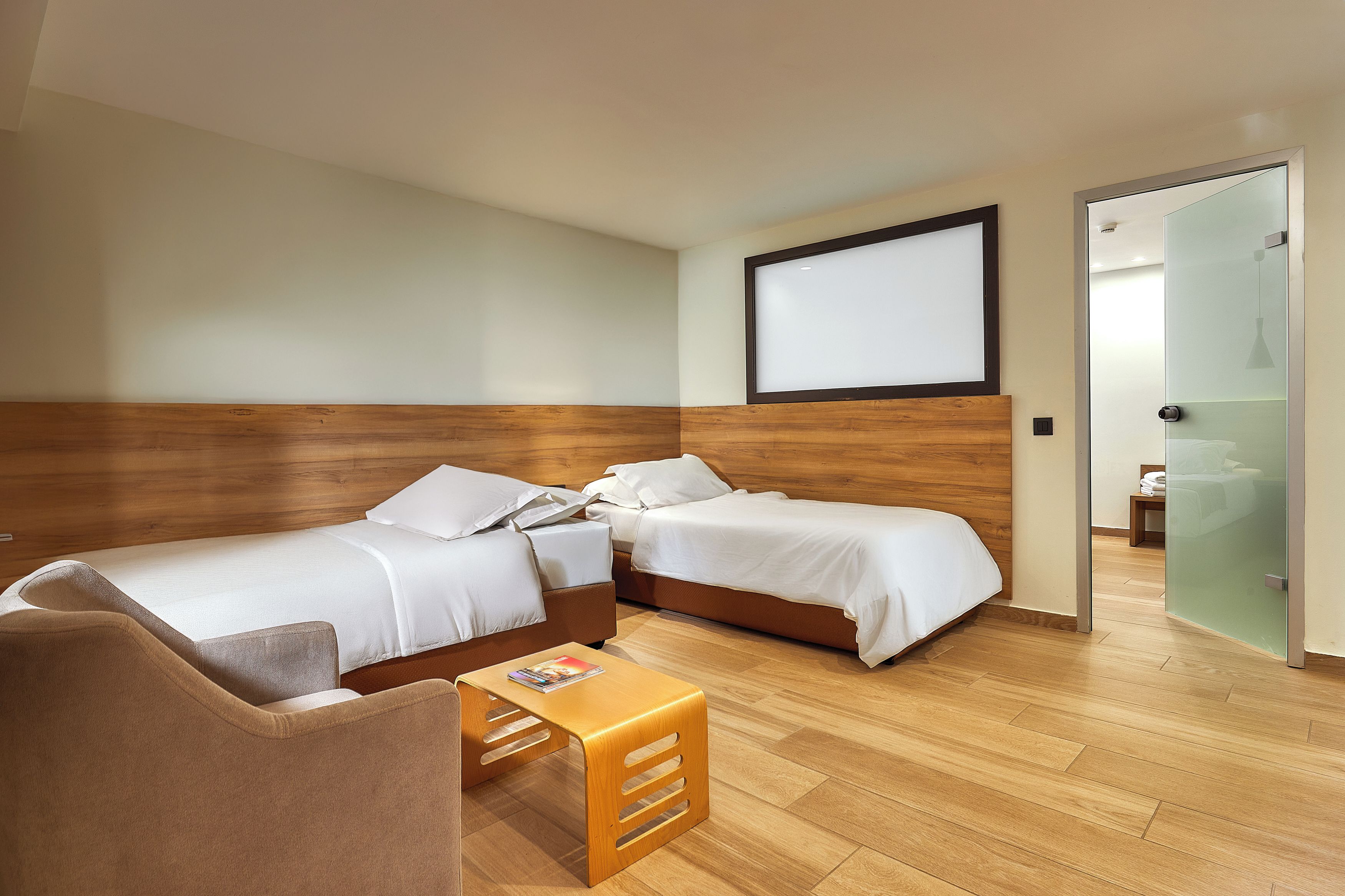 A hotel room with two beds, a wooden headboard, a small wooden table, and a glass door leading to a bathroom.