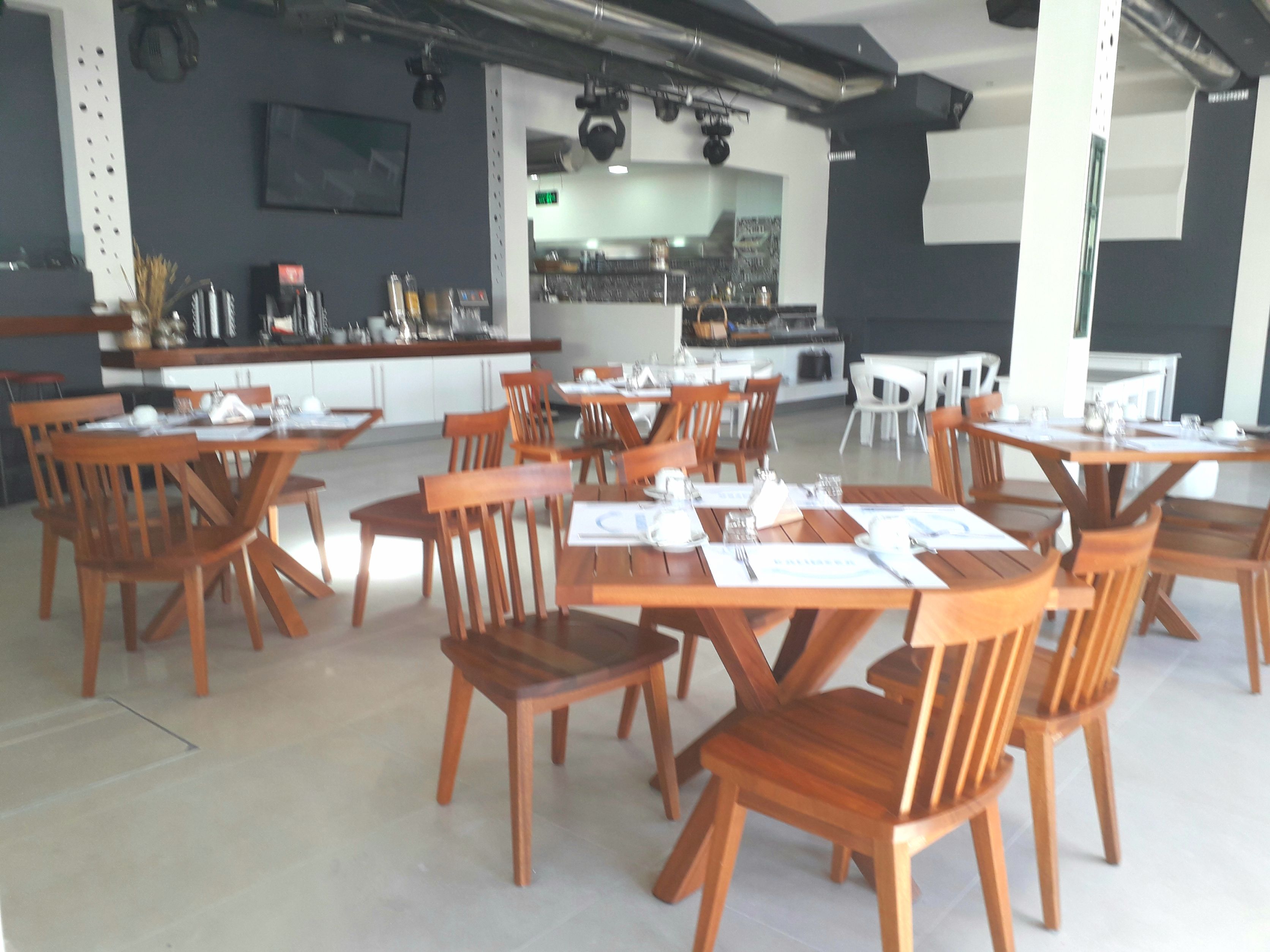 A restaurant with wooden tables and chairs, a kitchen in the background, and a modern interior design.