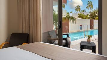 A hotel room with a bed, a chair, a small table with a plant, and a view of a swimming pool and palm trees.