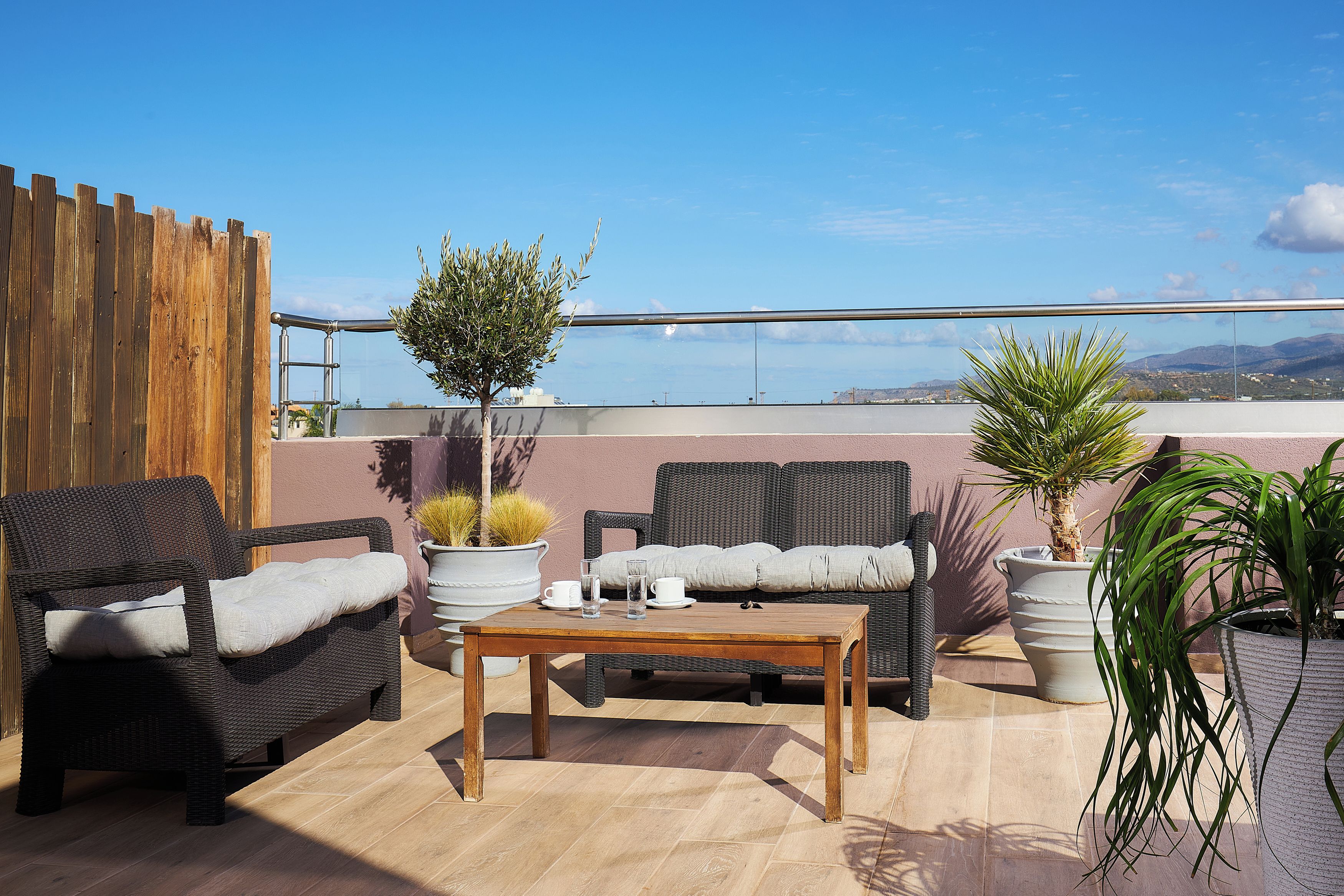 A rooftop terrace with wicker furniture, a wooden table, potted plants, and a view of the sea.