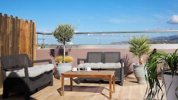 A rooftop terrace with wicker furniture, a wooden table, potted plants, and a view of the sea.