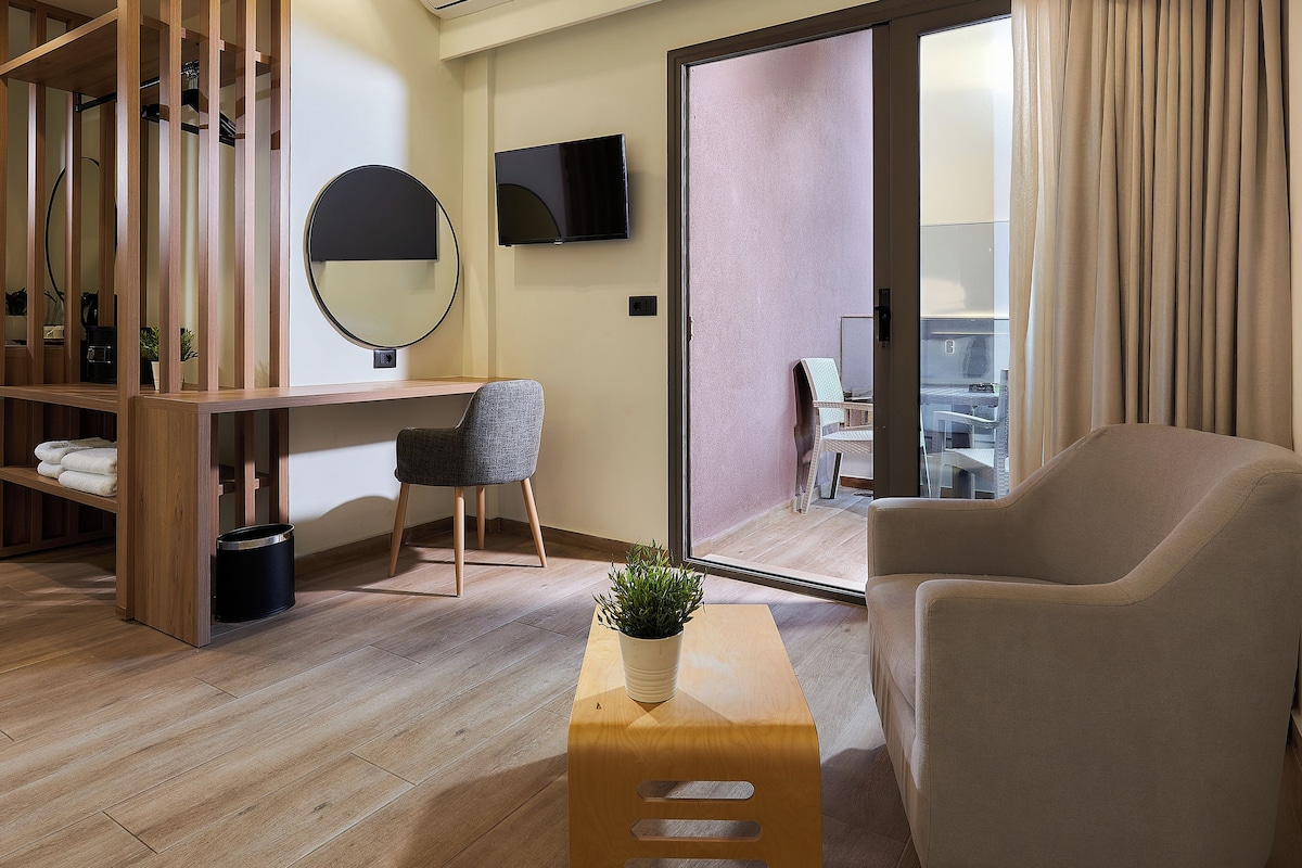 Moderni hotellihuone, jossa on puinen työpöytä, mukava nojatuoli, pieni pöytä, jossa on kasvi, pyöreä peili ja seinälle asennettu televisio.