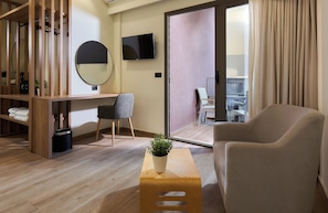 Una habitación de hotel moderna con un escritorio de madera, un sillón cómodo, una mesita con una planta, un espejo redondo y un televisor en la pared.