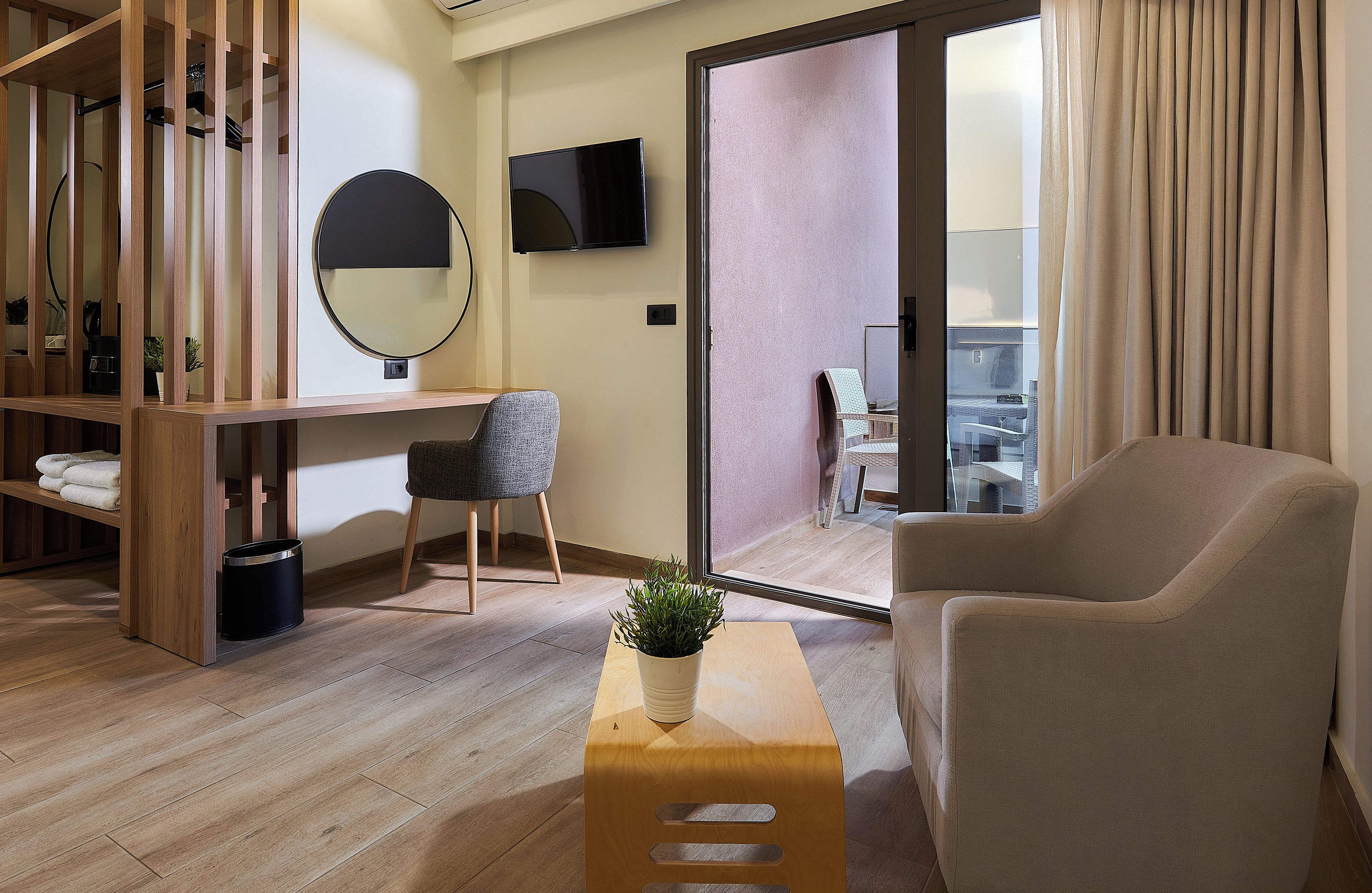 A modern hotel room with a wooden desk, a comfortable armchair, a small table with a plant, a round mirror, and a TV on the wall.