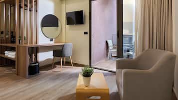 A modern hotel room with a wooden desk, a comfortable armchair, a small table with a plant, a round mirror, and a TV on the wall.