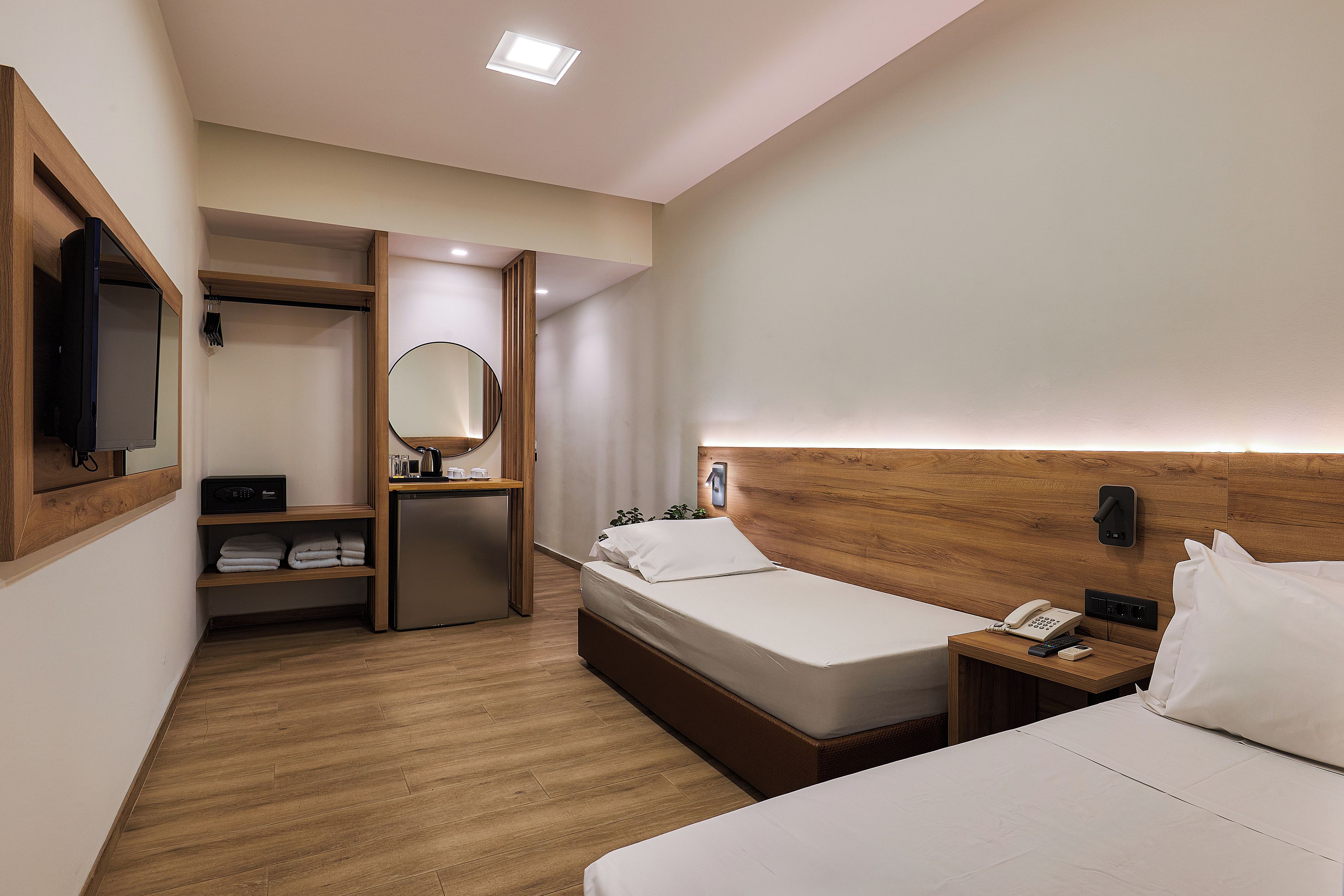 A hotel room with two beds, a wooden headboard, a bedside table with a phone and a lamp, a small desk with a mirror and a shelf with towels.