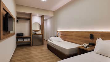 A hotel room with two beds, a wooden headboard, a bedside table with a phone and a lamp, a small desk with a mirror and a shelf with towels.