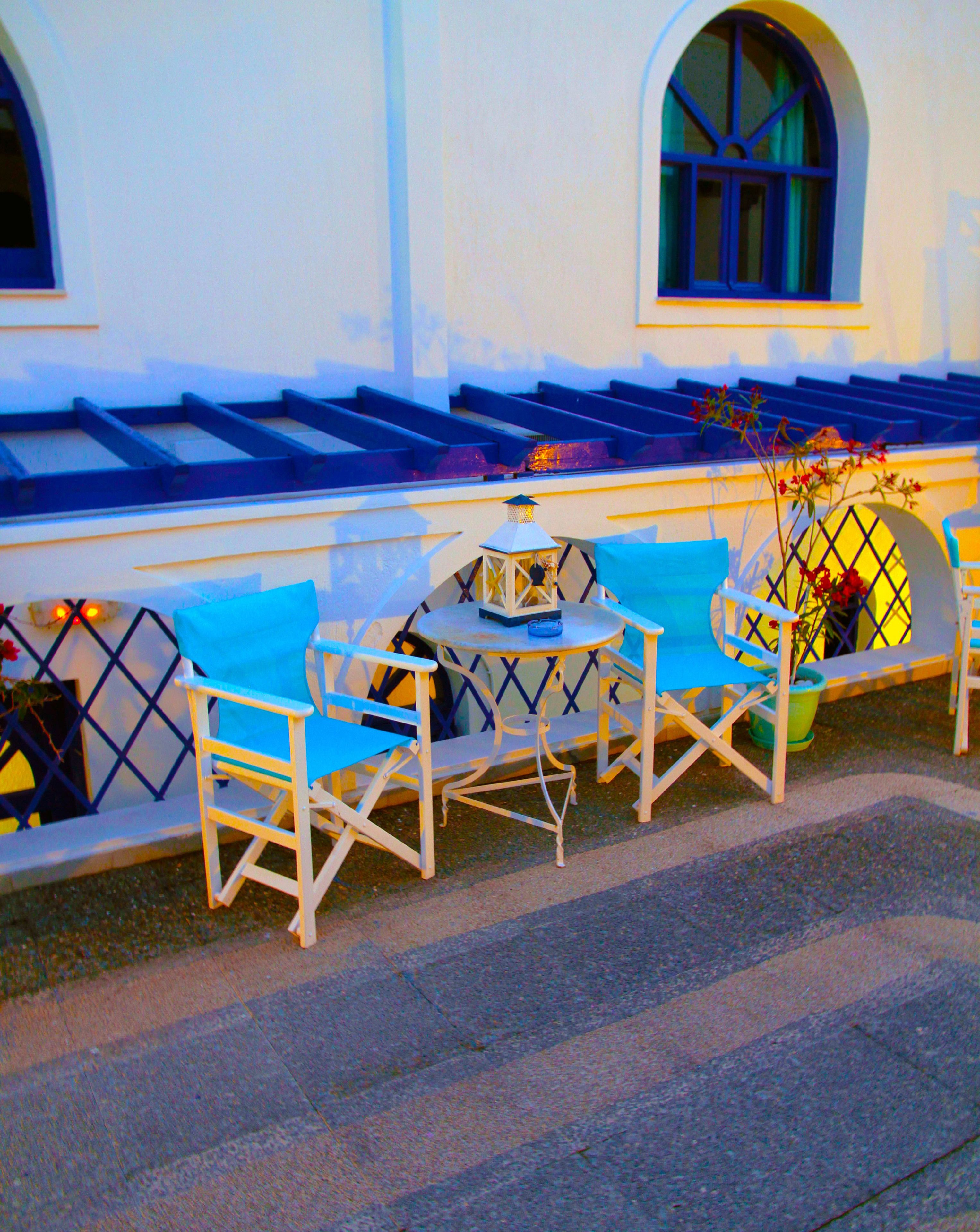 A seating area with blue cushioned chairs and a small table, set against a white building with blue accents and a window.