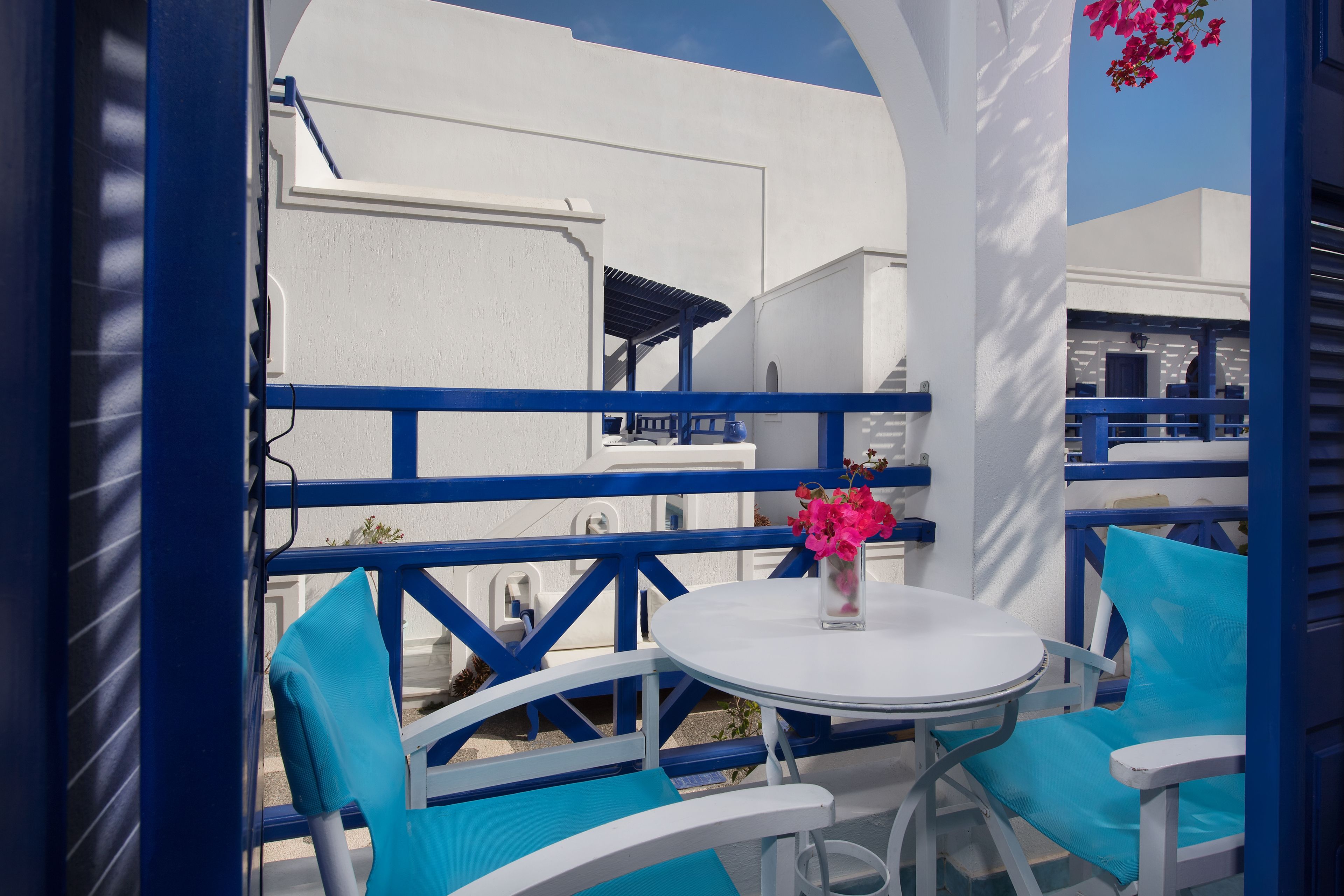 A white and blue outdoor seating area with a table and chairs, a vase with flowers, and a view of white buildings.