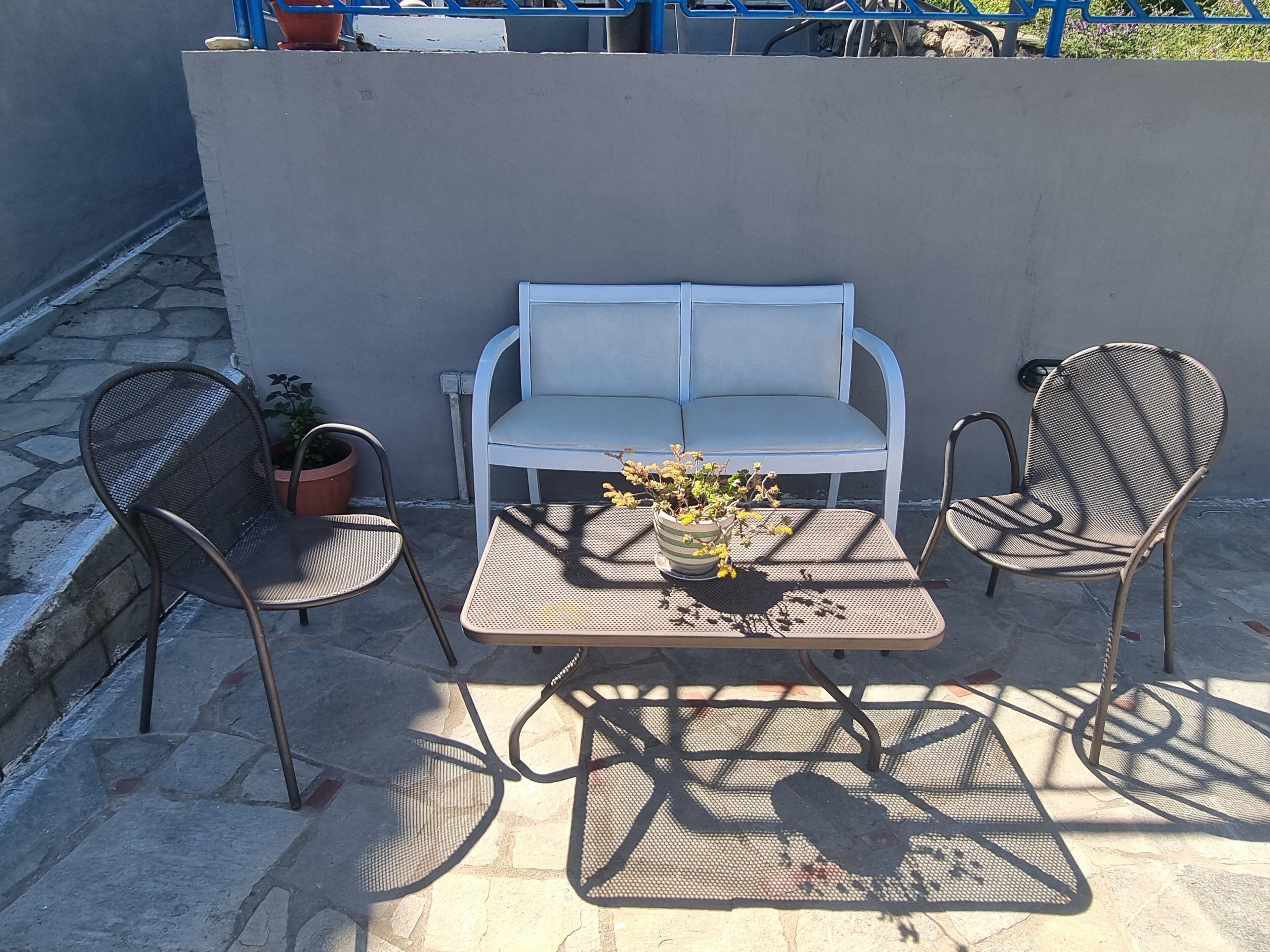 Een terras met een witte bank, twee metalen stoelen, een klein tafeltje met een bloempot en een blauwe balustrade op de achtergrond.