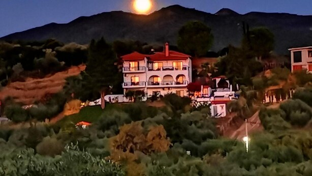A two-story building with a red roof and multiple balconies, surrounded by greenery and hills. - SartiVista (Sithonia)