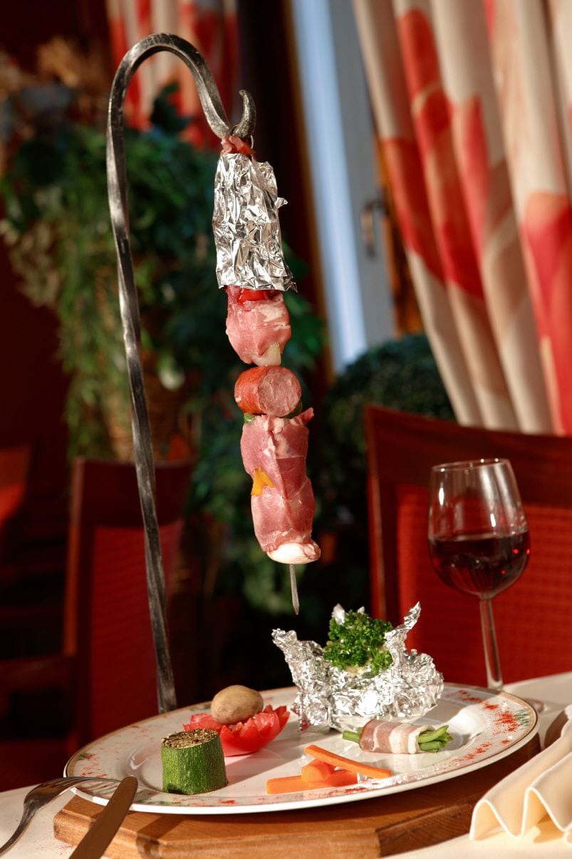 A plate with a skewer of meat, vegetables, and a small dish of greens, accompanied by a glass of red wine.