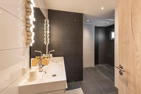 A modern bathroom with a white sink, two faucets, a mirror with lights, and dark tiled walls. - Hôtel - Restaurant Victor Hugo (Vianden)