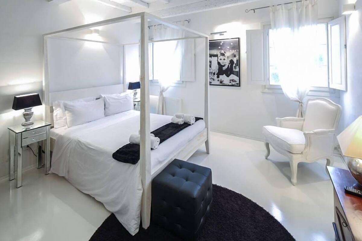Una cama con dosel con ropa blanca, una mesita de noche con lámpara, un reposapiés negro, un sillón blanco y un cuadro en la pared.