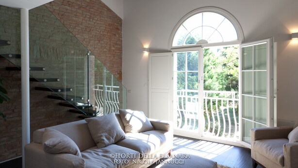 A modern living room with a glass staircase, a large arched window, and a view of greenery outside. - Villa Horti della Fasanara (Ferrara)
