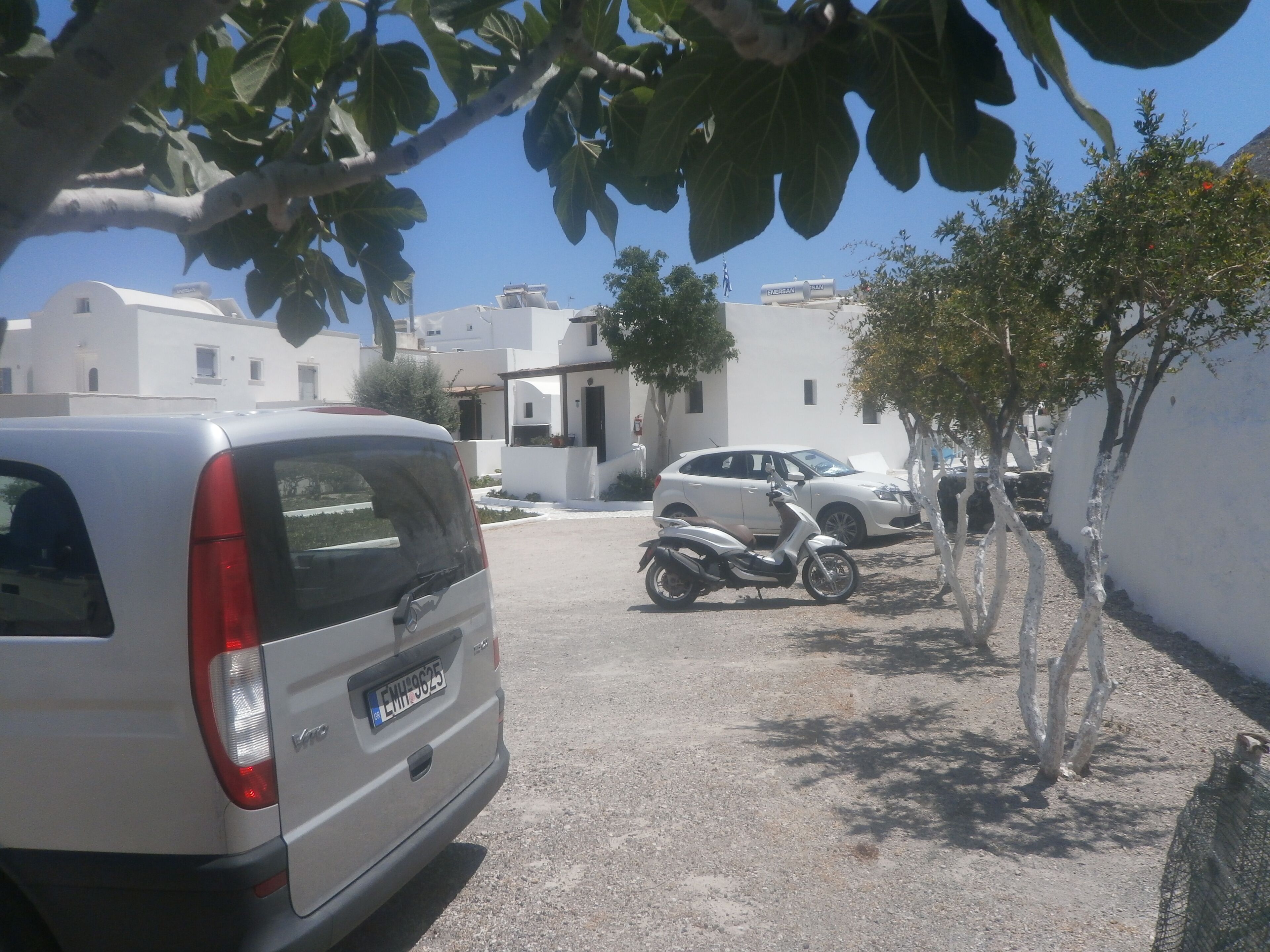 A parking area with a white van, a silver car, and a scooter. There are white buildings with flat roofs in the background.