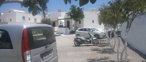 A parking area with a white van, a silver car, and a scooter. There are white buildings with flat roofs in the background.