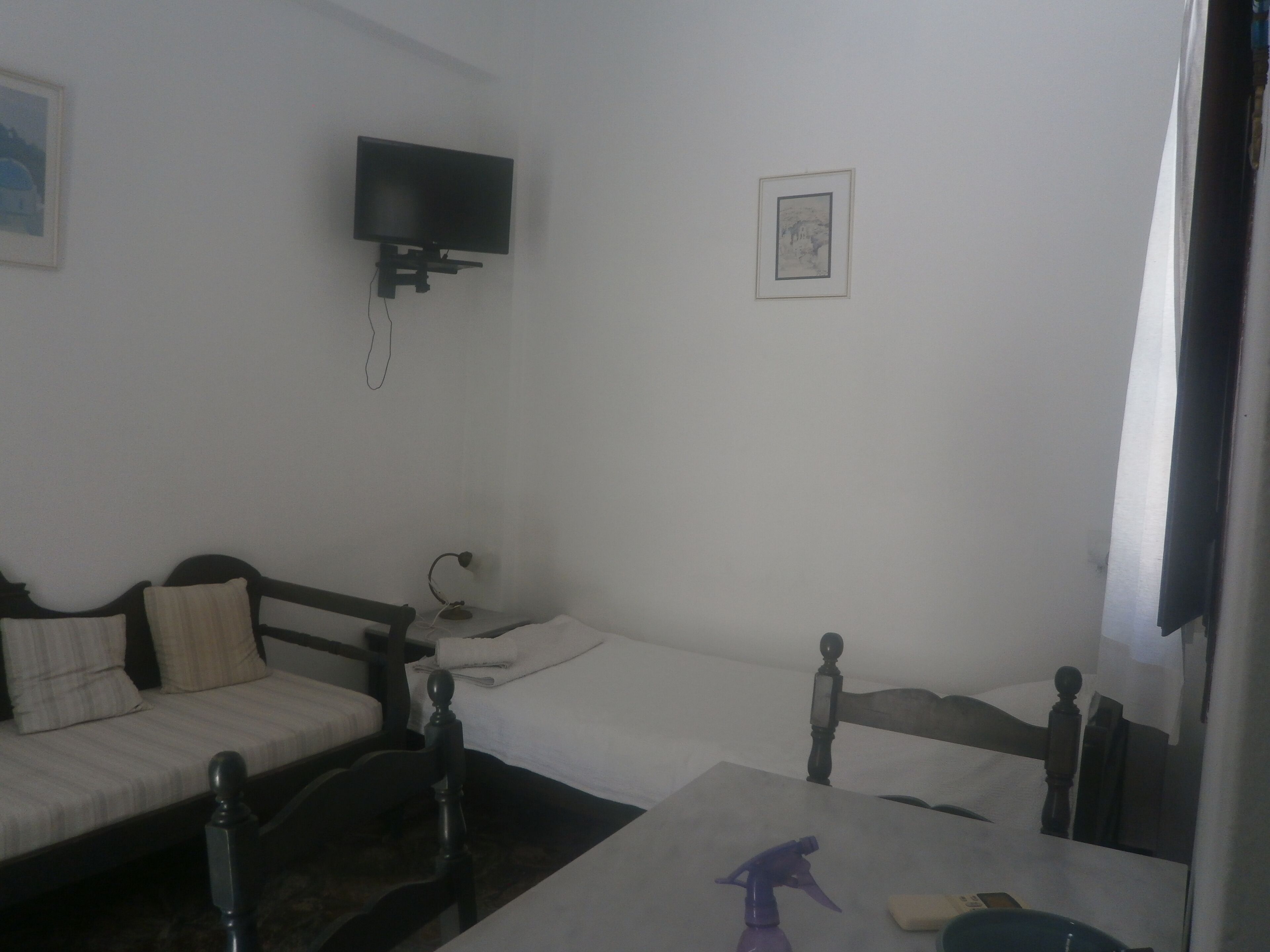 A room with a bed, a bedside table with a spray bottle, a small table with a soap bar, a chair, a sofa, a TV mounted on the wall, and a framed picture.