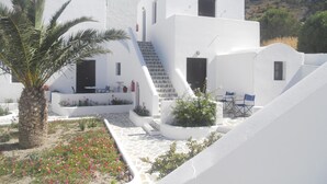 A white building with a staircase leading to a balcony, surrounded by a garden with flowers and a palm tree. - Villa Ostria (Santorini)