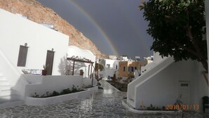A white building with a stone-paved courtyard, a mountain in the background, and a rainbow. - Villa Ostria (Santorini)