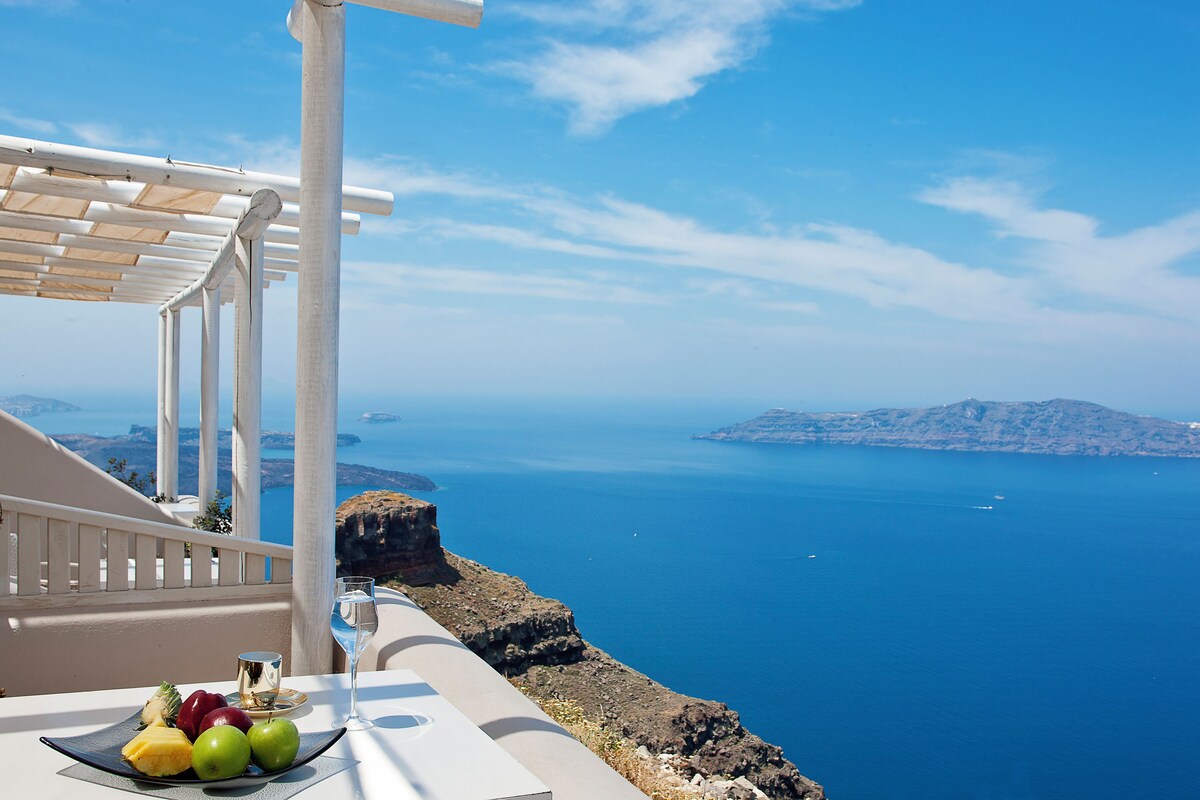 A balcony with a table set for a drink and fruit, overlooking a clear blue sea and distant islands.