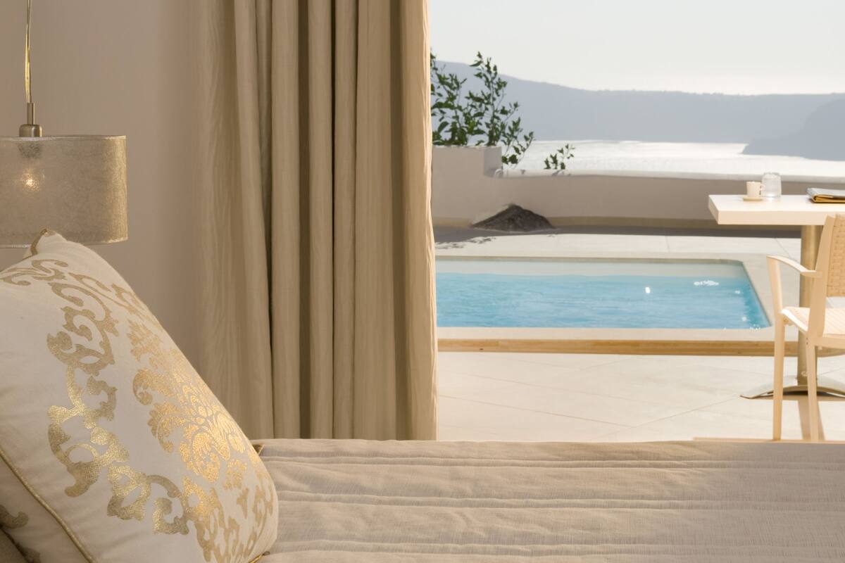 A bedroom with a bed, a nightstand, a lamp, a window with curtains, and a view of a pool and landscape.