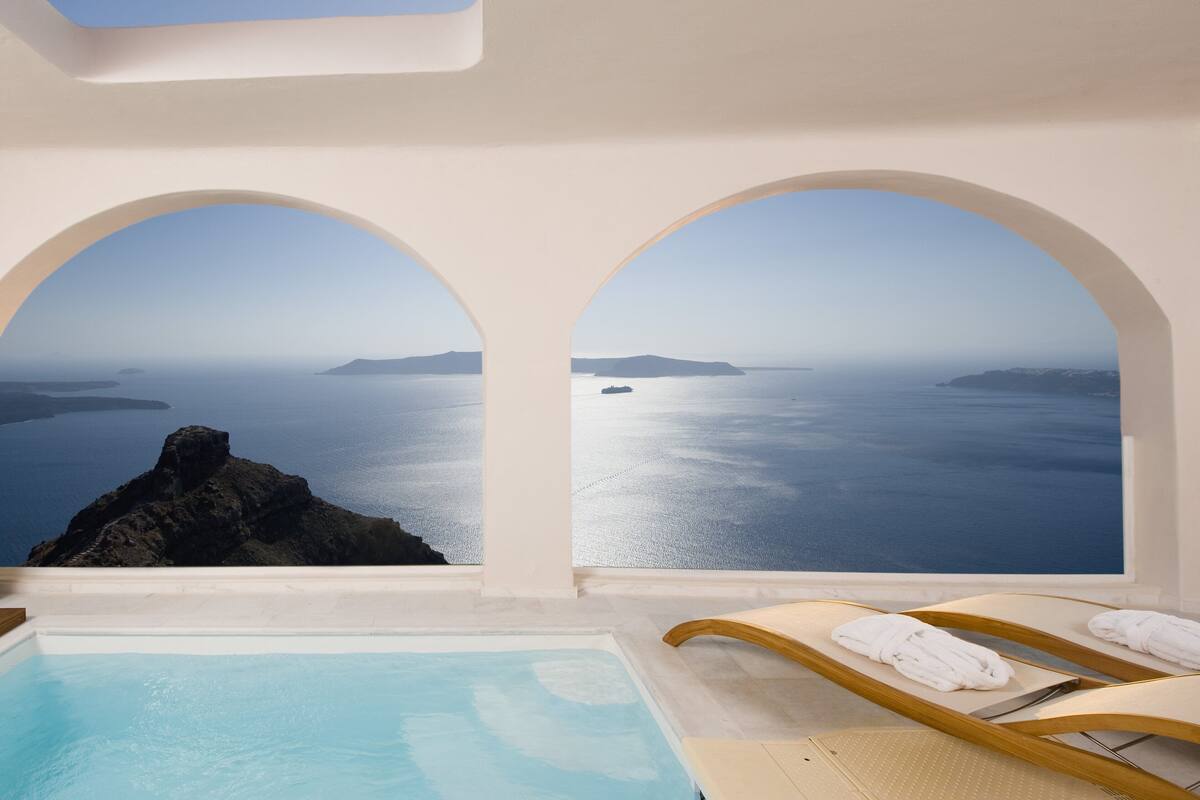 A pool area with two lounge chairs and towels, a view of the sea through arched windows, and a clear sky.