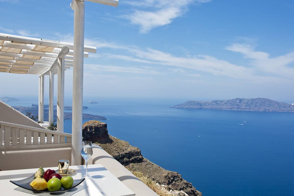 A balcony with a table set with fruit and a glass of water, overlooking a clear blue sea and distant islands.