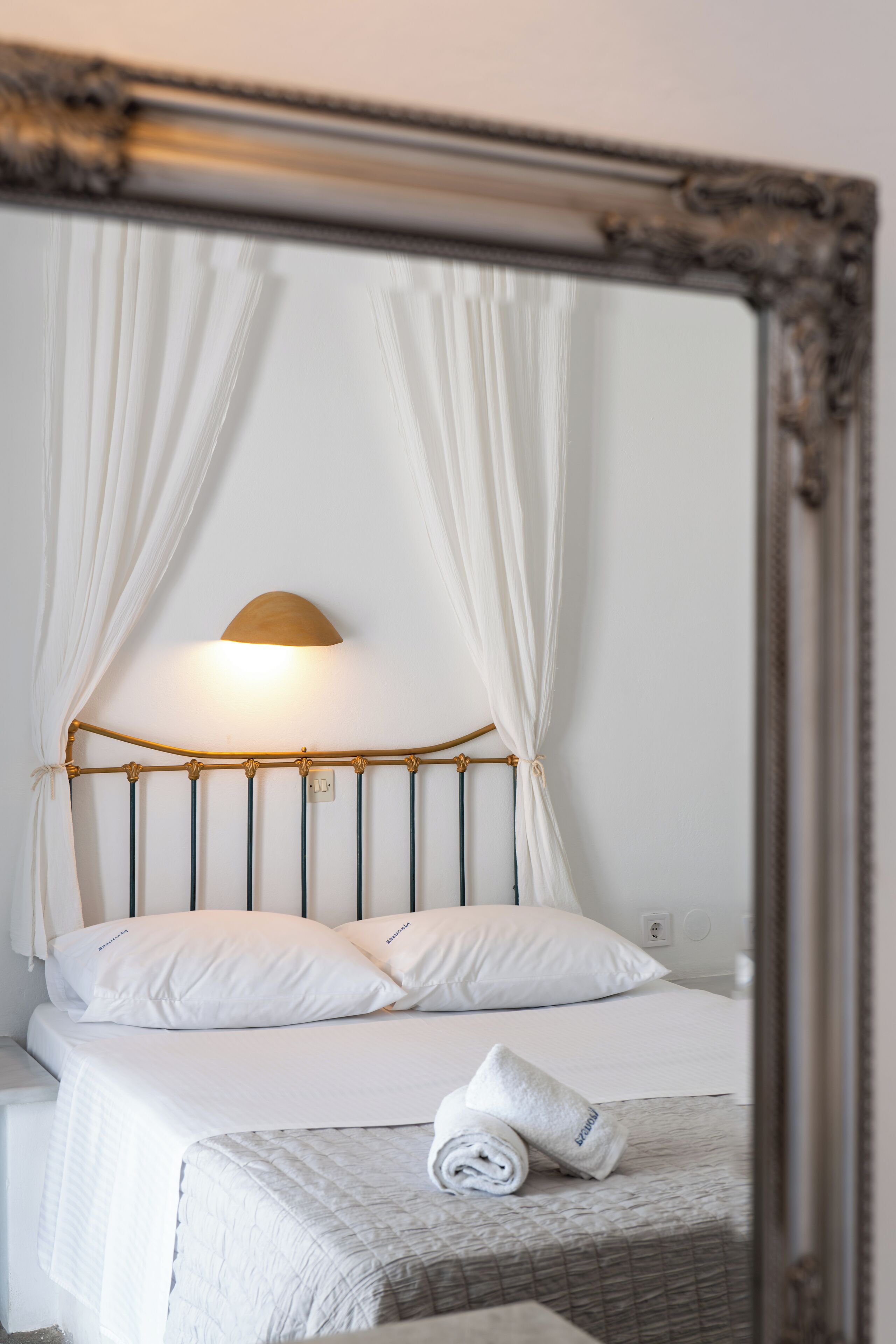 A neatly made bed with white linens, a headboard with a light fixture, and a mirror reflecting the room.