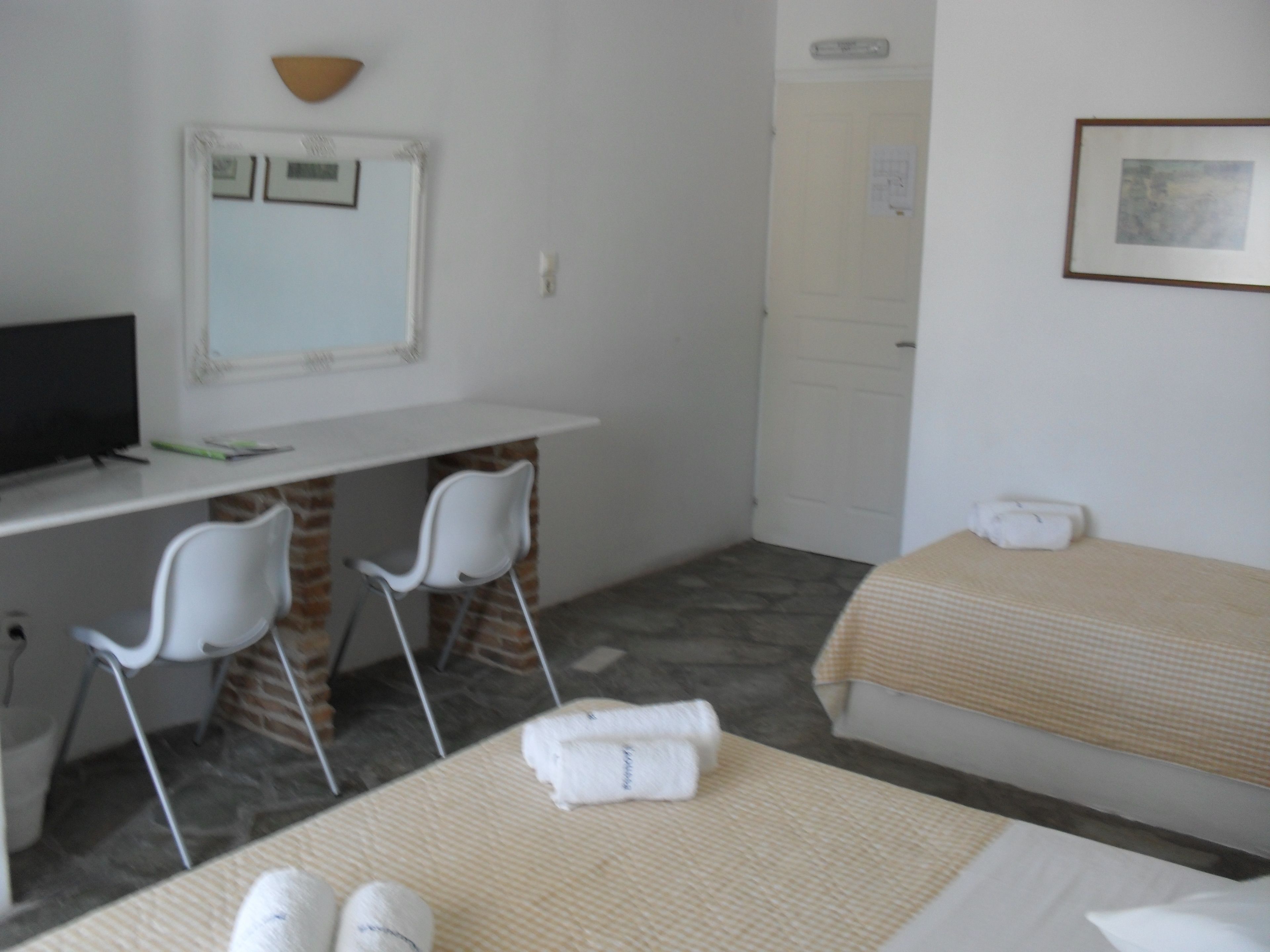 Une chambre d’hôtel avec un bureau, deux chaises blanches, un lit recouvert d’une couverture beige et un tableau encadré au mur.