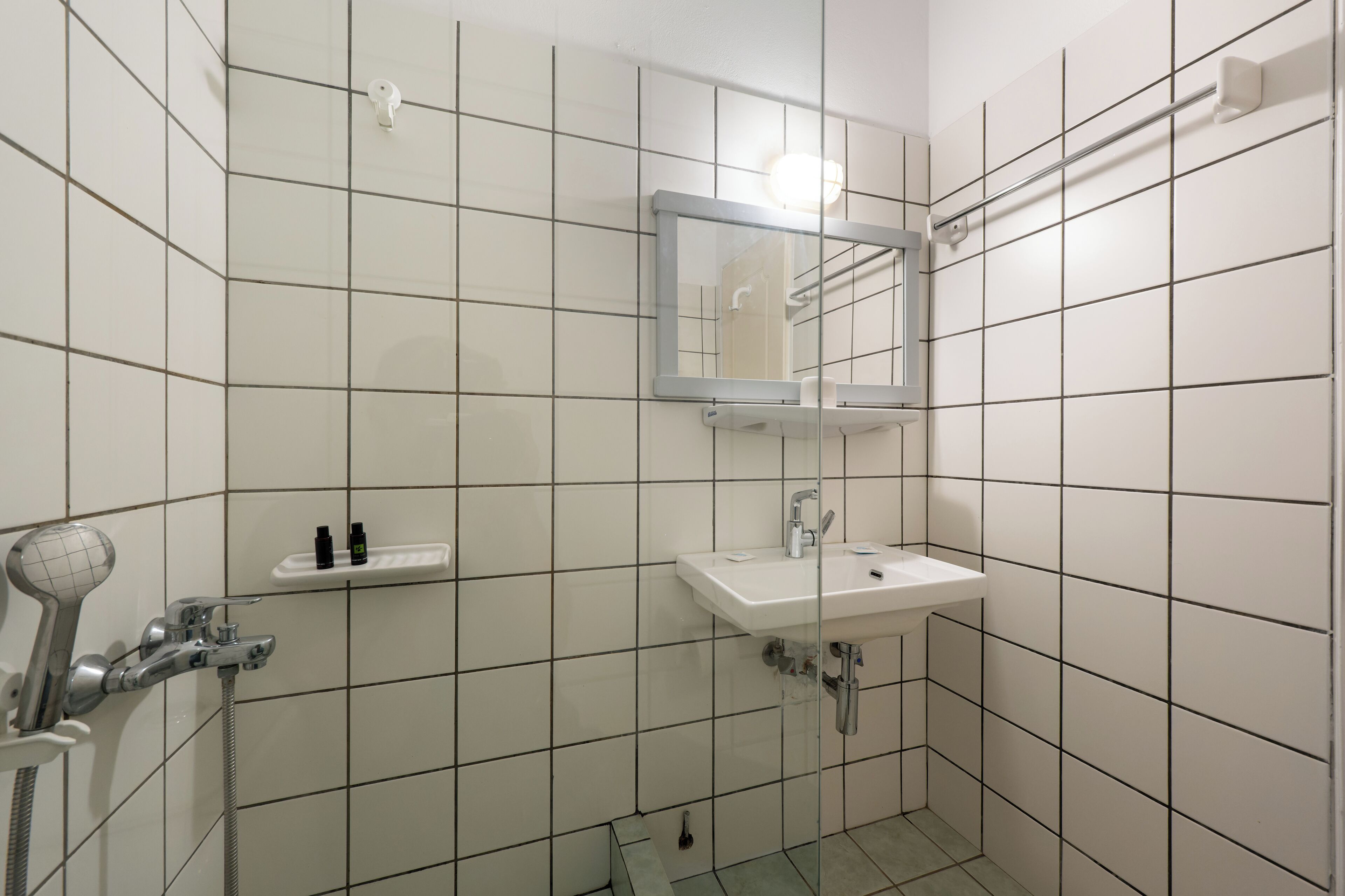 Et lille badeværelse med hvide flisevægge, en hvid vask, et spejl og et brusehoved.
