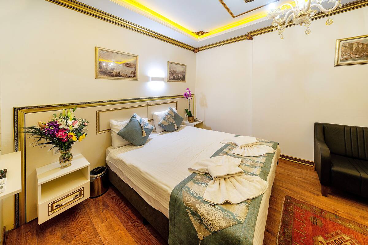 Une chambre d’hôtel avec un lit, une table de chevet, un vase de fleurs, un canapé en cuir noir et un lustre.