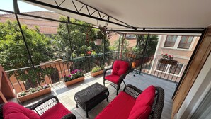 Ein Balkon mit Korbmöbeln, roten Kissen, Blumentöpfen und Blick auf benachbarte Gebäude.