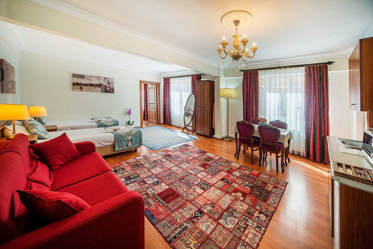 Une chambre d’hôtel avec un canapé rouge, un grand tapis, une table à manger avec des chaises et un lustre.