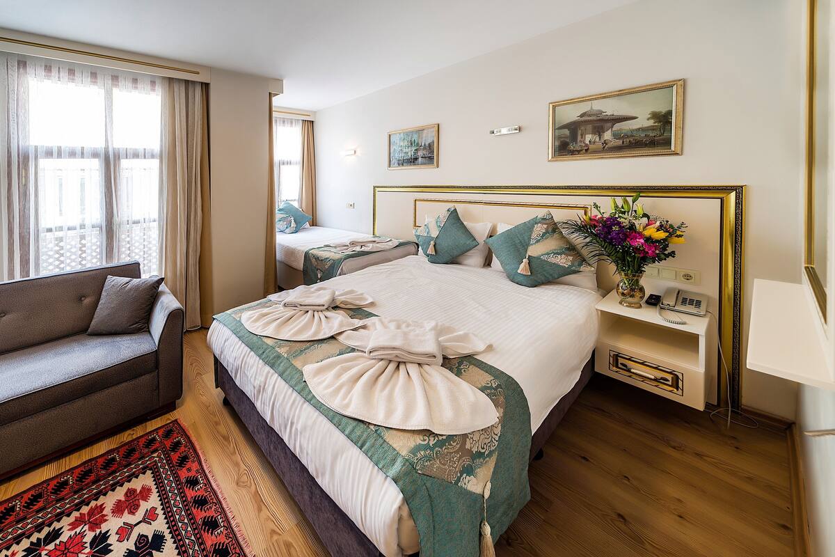 Une chambre d’hôtel avec un lit, un canapé, une table de chevet avec un téléphone, un vase de fleurs et un tableau au mur.