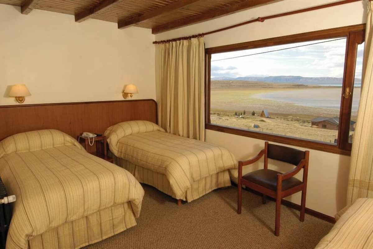Habitación de hotel con dos camas, una silla de madera, una mesita de noche y una ventana con vistas al paisaje.
