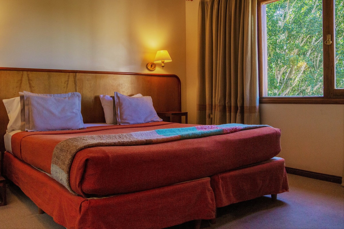 Una cama doble con una colcha roja, dos almohadas blancas y un cabecero de madera. Hay dos mesitas de noche con lámparas, una ventana con cortinas y vistas a árboles fuera.