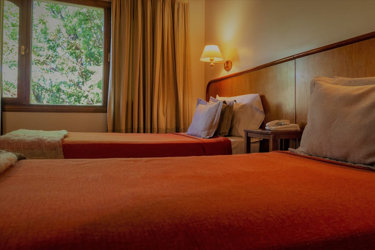 Una habitación de hotel con dos camas, una mesita de noche con lámpara y una ventana con cortinas.