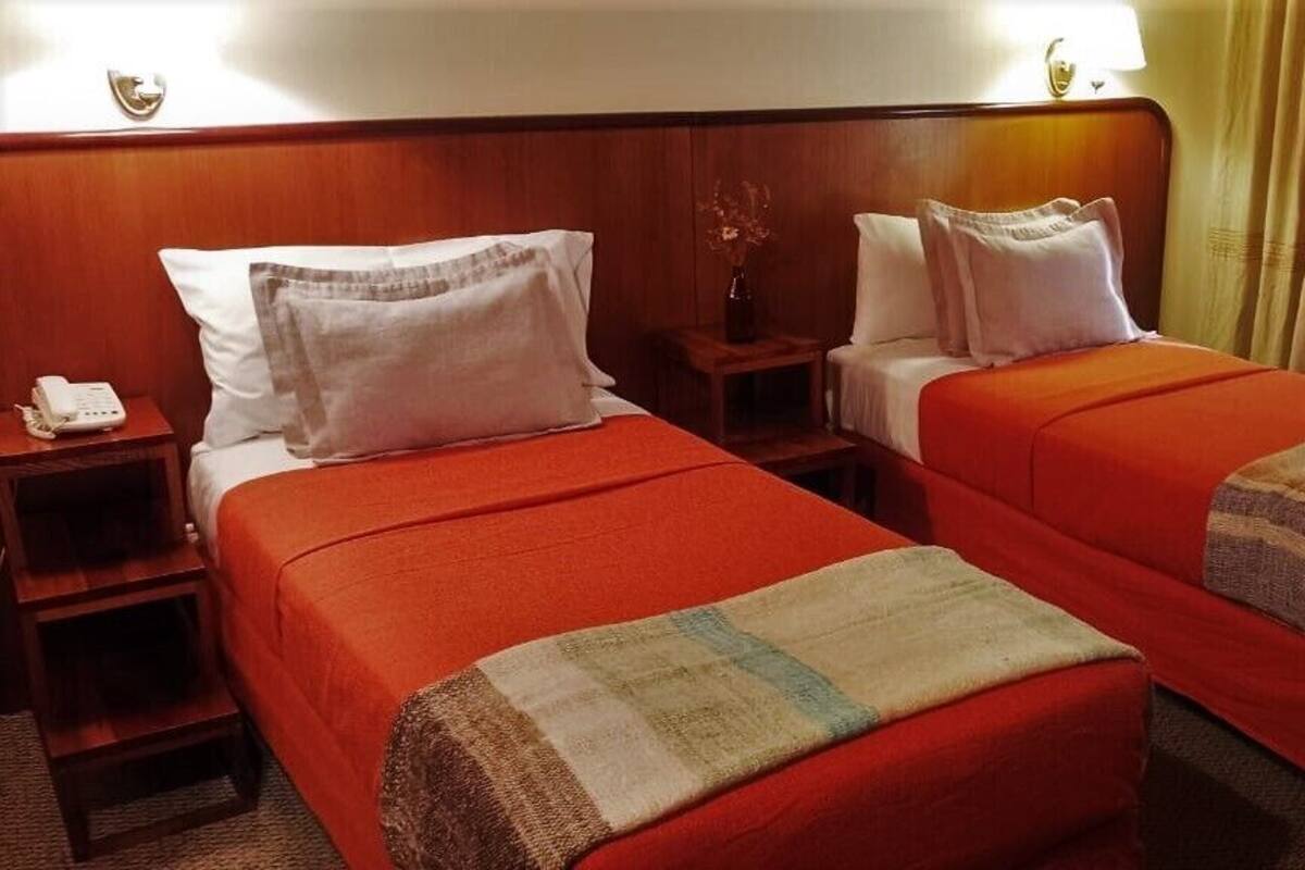 Habitación de hotel con dos camas, cada una con una colcha roja y almohadas blancas. Se observa un mesita de noche con una lámpara y un jarrón con flores.