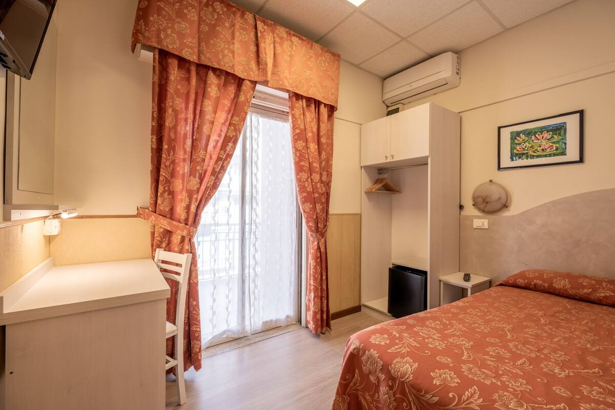 Una habitación de hotel con cama, escritorio, silla, armario y ventana con cortinas.
