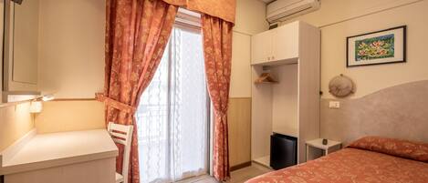 Una habitación de hotel con cama, escritorio, silla, armario y ventana con cortinas.